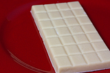 A photograph of a vegan white chocolate, scored in squares bar. It is placed on a red shiny circular surface. The background is blurred.