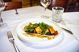 close-up on a fine dining table with white tablecloth and crystal wine and water glasses, and a white plate featuring a dish of greens and tofu with sauce, elegantly plated