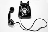 An image of an all black rotary phone with the earpiece laid out on the left and the cord slightly tangled beneath it on a greyish background.