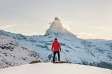 A man in a red coat on a snowy mountaintop looking at a sunrise.