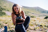 A happy woman sits in a field eating from a bowl.