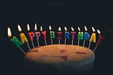 A white cake with rainbow candles, spelling out happy birthday on a black background