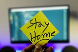 A hand holding a yellow sticker with the lettering: Stay Home.