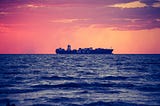 A cargo ship on the ocean, sat against a canvas sky of red and orange