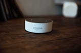 The image features an Amazon Echo Dot (3rd Generation) on a wooden surface. The device is compact and circular, primarily white with a gray top that includes speaker holes. In the background, there’s a slightly blurred white object, enhancing the focus on the Echo Dot.