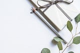 envelope tied with a thin grey pink ribbon, silver pencil alongside, a sprig of greenery lying along the bottom, white background.