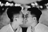 Two Asian people with short hair and glasses are looking at each other, smiling, very close together. They look like maybe they might kiss. Their gender is ambiguous.