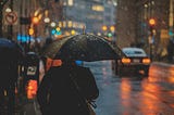 it’s evening on a city street, it’s raining, there’s a person walking with an umbrella and cars driving with their lights on