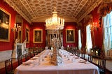Ornate dinner room with white statues and artwork hung on the surrounding red-orange walls with a centered long table for seating 16+ people
