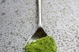 A metal spoon sits on a countertop, holding some sort of green powder—possibly matcha.