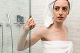 A white woman standing in a white-tiled shower. She has her hair wrapped in a white towel on top of her head, and her body is wrapped in another white towel. She is gripping the glass wall of the shower with her right hand.