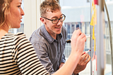 Two colleagues work together at a whiteboard, writing on several posted sticky notes.