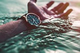 a man’s forearm and hand rest on top of the ocean with his watch-face upturned towards the sky