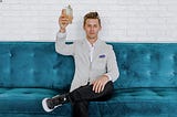 A man in a blazer on a blue sofa, proudly holding up a cocktail.