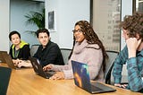 4 adults of different genders and races working together at a wooden table with laptops in from of them.