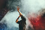 A music artist on stage reaching up with one hand while they sing. The crowd is shining lights in the background, and a white smoke envelopes the scene.