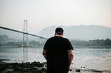 Man standing at a river’s edge pondering, bridge and mountains in the distance