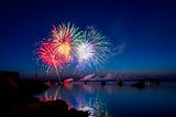 Multi-colored fireworks exploding over a river, against a deep blue sky.