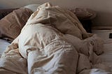 Someone wrapped up in tan sheets. They appear to be so cozy that they've even covered up their heads. Its summer, there is sunlight coming to the window shining onto the blankets.