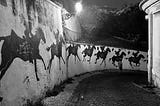 A nighttime view of a street in Lisbon, the cobbled calçadas gleaming in teh streetlight, the wall painted with a horse and rider galloping further and further away around the bend.