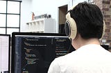 A young male programmer sits in front of a computer monitor displaying code.