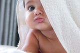 A colour photo of a pouting brown-eyed baby with a white towel partly over its head.