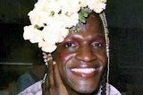 Portrait of Marsha P. Johnson smiling with white flowers in her hair and mauve makeup