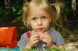 A young girl looking as as she holds a slice of salami in her hands, close to her mouth