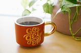 A coffee cup sits on a desk in front of a potted house plant. The side of the cup reads ‘Go get ‘em’.
