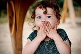 A white girl about age four with light brown curly hair holds her hands to her face in a surprised expression. She has big blue eyes and wears a green and white polka dot T-shirt. She is in focus with a few wooden structures out of focus in the background.