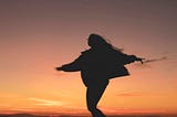 The silhouette of a person with long hair and glasses dancing on a sunset beach.