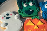 Decorated Halloween cookies shaped like a ghost, Frankenstein’s monster, and a jack-o’-lantern on a white plate — symbolizing how fear is sweetened and made harmless.