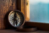 An antique brass compass with a white face sits on a wood frame looking out a wondow to blue sky