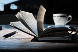 An open journal and pen sit on a desk with a teacup in the background.