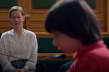 An image shows a snippet from the movie Anatomie d’une Chute. Sandra Huller as Sandra Voyter sits in court wearing a white top and black pants. Milo Machado-Graner, who plays Daniel in the film, is in the forefront.