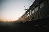 Photo of a passenger train heading off into a foggy horizon. The colors are drab and the image is gloomy and mysterious. The train might be derelict. Photo by Andrey Svistunov on Unsplash.