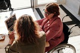 A women in a wheelchair looking at a computer screen with another women sitting next to her.