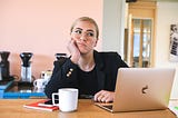 Woman wearing glasses, sitting at table with laptop and coffee mug, looking lost.
