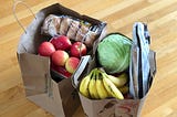 Two brown shopping bags filled with food such as bananas, red apples, bagels, iceberg lettuce and bread.