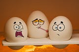 Three white eggs with cartoon faces expressing different emotions — one frightened, one sick with a thermometer, and one worried — sitting together on a shelf under warm lighting, symbolizing fear, anxiety, and vulnerability in a humorous way.