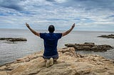 A person kneels on the beach, their back to the viewer, facing the ocean, with their arms raised to the sky.