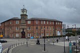 View across a wide pavement and road towards an old brick three-storey building on a corner. A stone clocktower that is taller than the rest of the building is set at the corner, which wings in brick either side