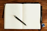 A blank journal laying open on a wood surface with a pen resting on top of the pages.