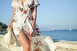 Girl wearing cream dress with floral print, carrying a sun hat by the sea