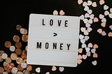Sign saying, “Love > Money” on top of a pile of coins.