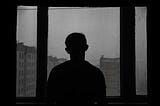 The silhouette of a man looking out of a window at several plain concrete buildings. The sky is overcast and everything is gray.