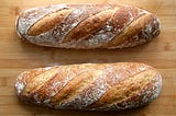 This is a photo of two loaves of unsliced bread. They may be homemade or from a bakery and they have flour visible on top.