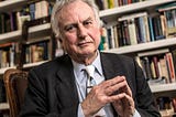Richard Dawkins sitting on a chair with a bookshelf in the background.