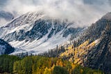A rugged snow-dusted mountain arrayed in swirling clouds towers over a sweeping valley with evergreen trees and aspen changing from green to gold.