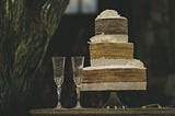 Two empty champagne flutes and a three layer wedding cake with brown and white frosting on a table